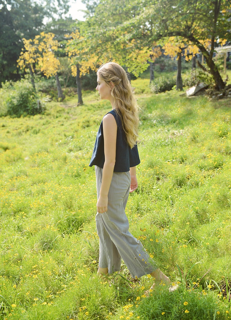 May include: A woman with long, wavy blonde hair walks through a field of green grass and yellow wildflowers. She is wearing a dark blue sleeveless top and gray pants.