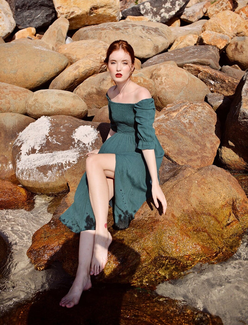 May include: A woman wearing a teal green off-the-shoulder dress sits on a large rock by the water. She has red hair and is looking directly at the camera.