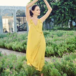 May include: A woman wearing a flowing, yellow wrap dress with ruffled trim, standing in a garden. The dress has a V-neck and a wrap design. She is wearing brown ankle boots. The background includes a building with arched windows and lush greenery.