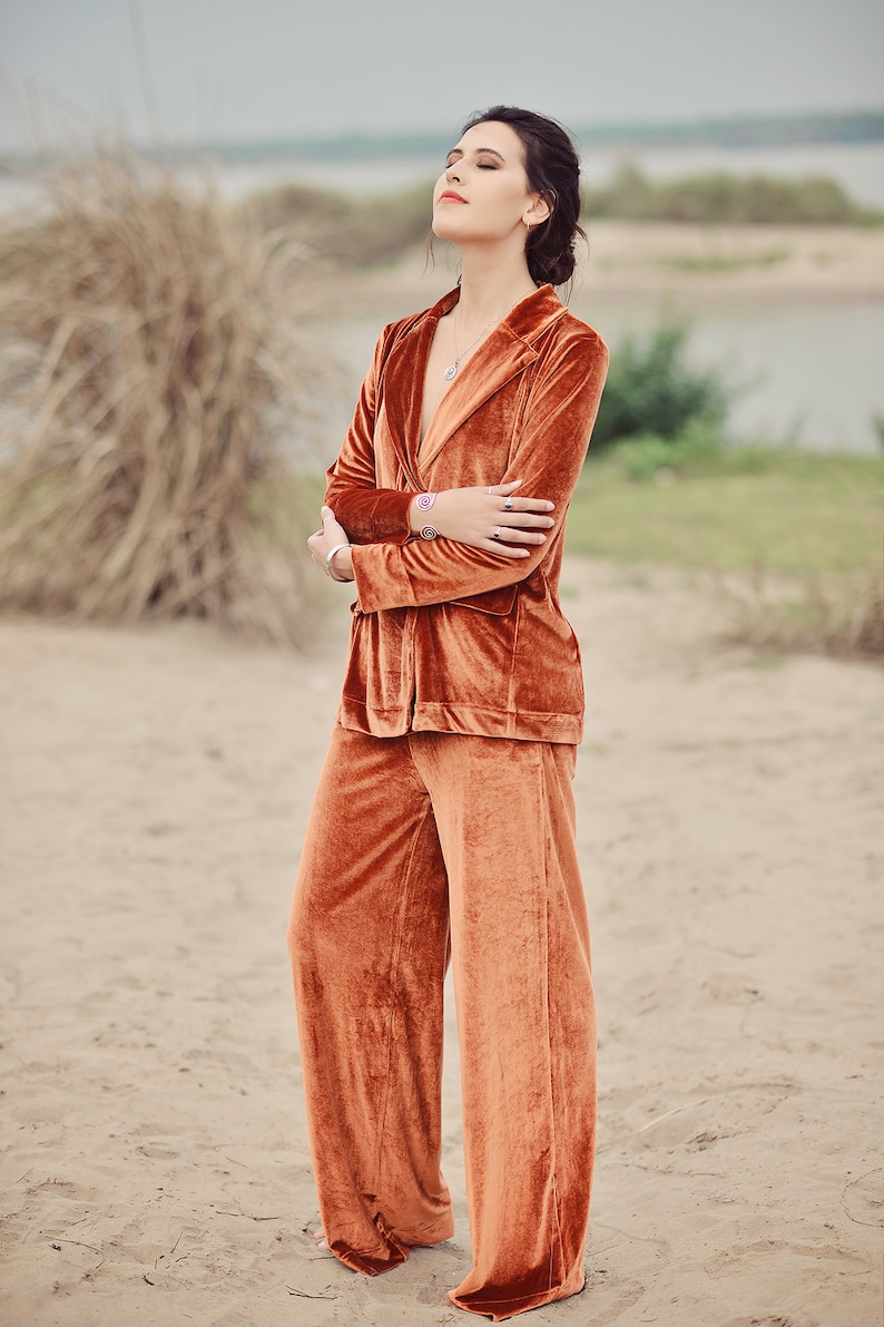 May include: A woman wearing a rust-colored velvet pantsuit with a deep V-neckline. She is standing with her arms crossed and her eyes closed.