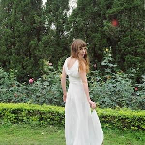 May include: A woman in a white sleeveless gown stands in a garden with green bushes and pink flowers. She is looking over her shoulder and holding the skirt of her dress.