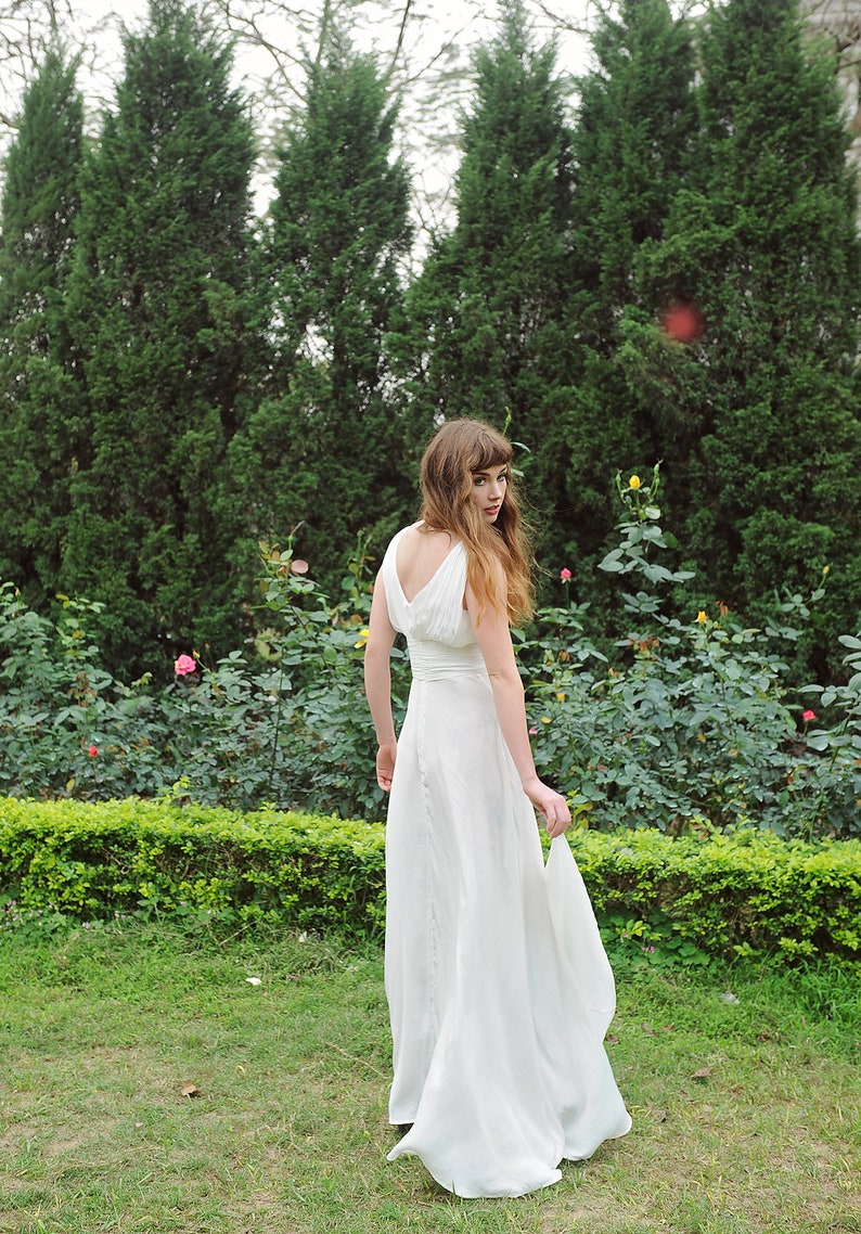 May include: A woman in a white sleeveless gown stands in a garden with green bushes and pink flowers. She is looking over her shoulder and holding the skirt of her dress.