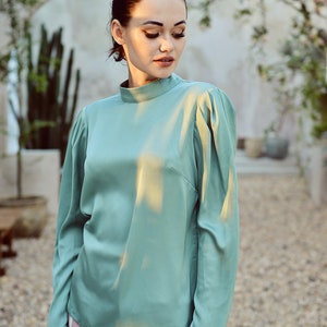 May include: A woman wearing a light green, long-sleeved blouse with a high neck and a pink skirt.