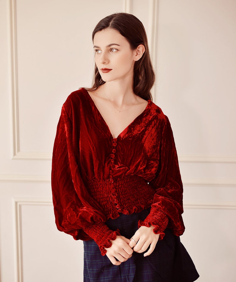 May include: A red velvet blouse with a V-neckline, long sleeves, and a button-up front. The blouse has a fitted bodice and a gathered waist.