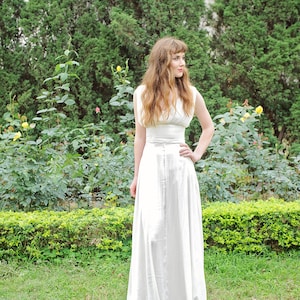 May include: A woman in a white sleeveless dress stands in a garden with green bushes and yellow flowers. She is looking to the right of the frame.