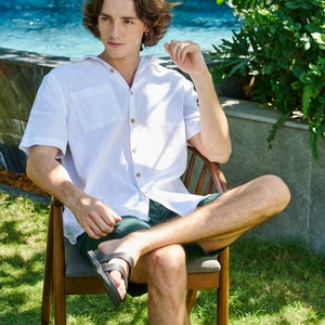 May include: A man wearing a white linen shirt with a pocket and short sleeves sits in a wooden chair with green shorts and black sandals. He is sitting in a grassy area with a pool in the background.