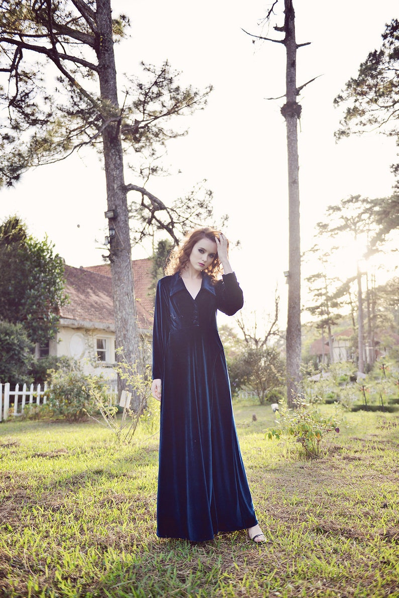 May include: A long, dark teal velvet dress with long sleeves and a button-down front. The dress is worn outdoors in a grassy area with trees and a house in the background. The dress has a classic, elegant style.