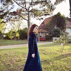 May include: A long, dark teal velvet dress with long sleeves. The dress is worn outdoors in a grassy area with trees and a house in the background. The dress has a high neckline and a flowing skirt.