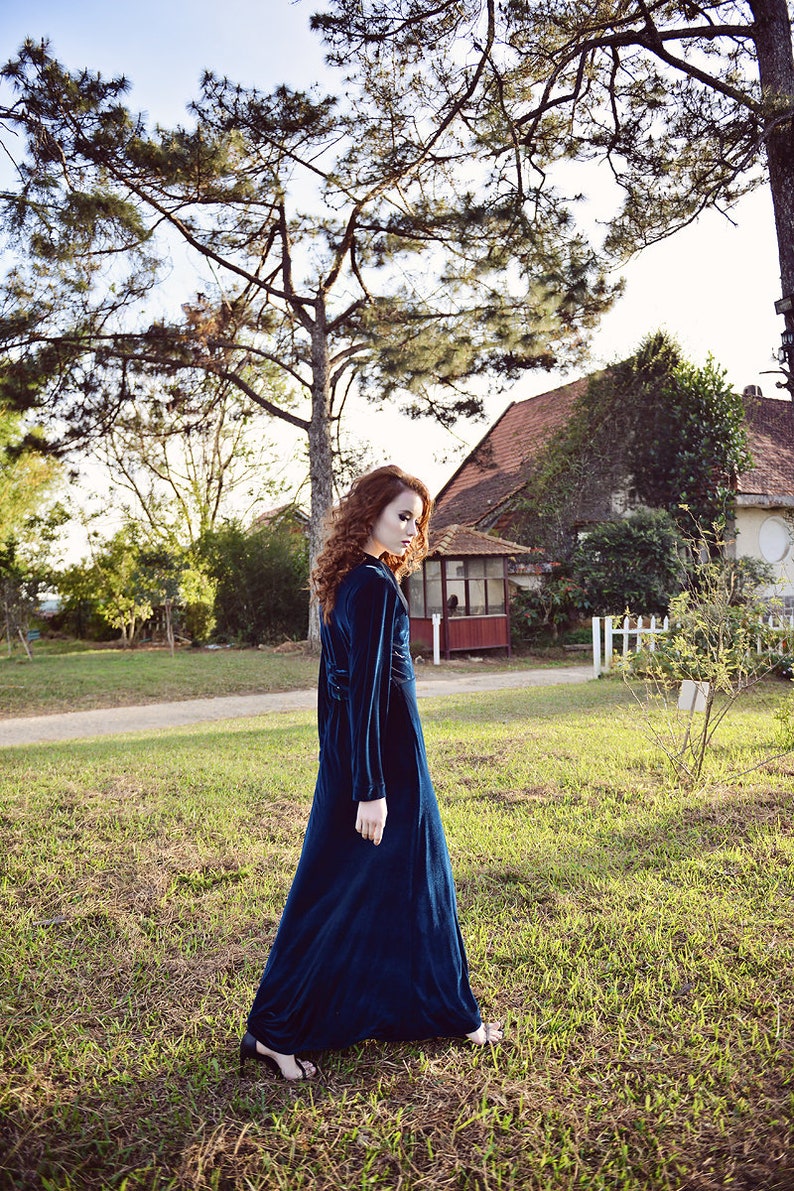 May include: A long, dark teal velvet dress with long sleeves. The dress is worn outdoors in a grassy area with trees and a house in the background. The dress has a high neckline and a flowing skirt.