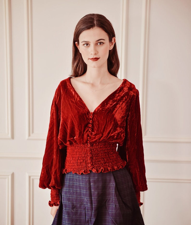 May include: A red velvet blouse with a v-neck, long sleeves, and a fitted waist. The blouse has a button-up front and a ruffled hem.