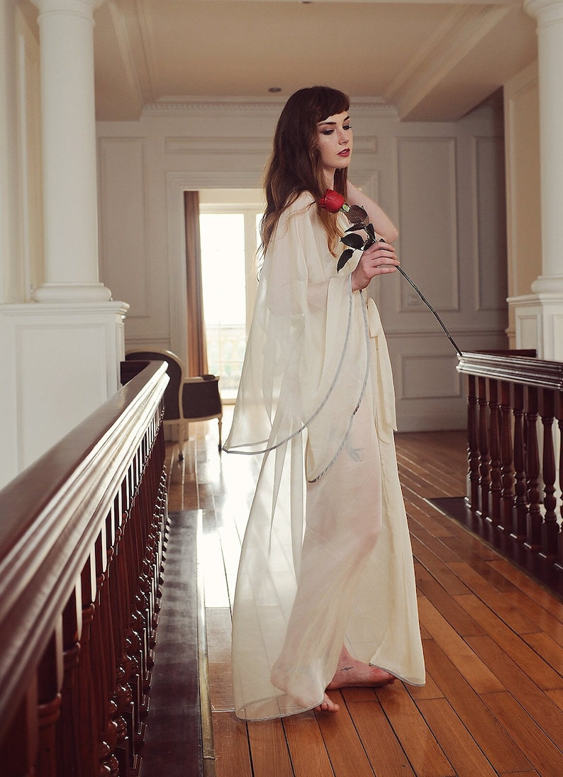 May include: A woman in a white sheer robe with a single red rose in her hand stands on a wooden floor in a hallway with a wooden railing.