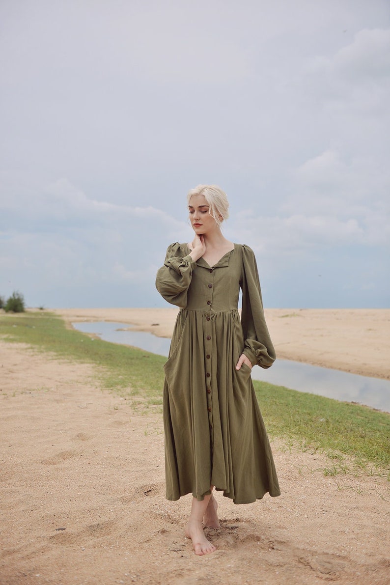 May include: A woman wearing a long-sleeved, olive green button-down dress with a gathered skirt and puffed sleeves. She is standing on a sandy beach with a small stream of water in the background.