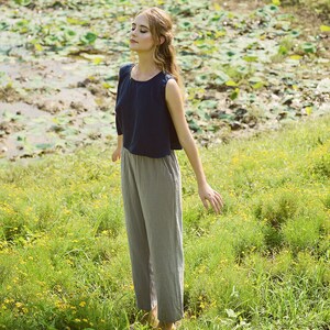 May include: A woman wearing a navy blue sleeveless top and gray pants stands in a field of green grass with yellow flowers. She has her eyes closed and her arms are outstretched.