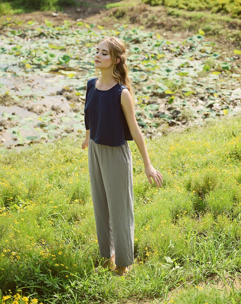 May include: A woman wearing a navy blue sleeveless top and gray pants stands in a field of green grass with yellow flowers. She has her eyes closed and her arms are outstretched.