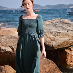 May include: A woman wearing a dark green, square-neck midi dress with puffed sleeves stands on a rocky beach. The dress has a smocked bodice and a flowing skirt.
