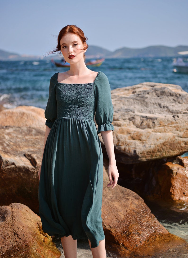May include: A woman wearing a dark green, square-neck midi dress with puffed sleeves stands on a rocky beach. The dress has a smocked bodice and a flowing skirt.