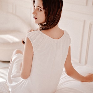 May include: A woman wearing a white, sleeveless nightgown with a gathered neckline sits on a white bed.