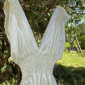 May include: Ivory-colored dress with a V-neck and short sleeves. The bodice features intricate smocking detail, creating a textured effect. The dress is made of a flowing, lightweight fabric, perfect for warm weather. The dress is hanging on a tree branch.