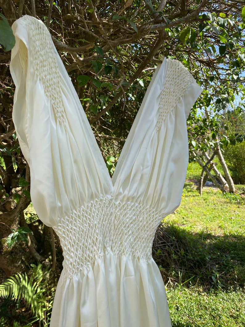 May include: Ivory-colored dress with a V-neck and short sleeves. The bodice features intricate smocking detail, creating a textured effect. The dress is made of a flowing, lightweight fabric, perfect for warm weather. The dress is hanging on a tree branch.