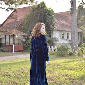 May include: A long, dark teal velvet dress with long sleeves and a cinched waist. The dress is worn outdoors in a grassy area with a house in the background. The dress has a high collar and a flowing skirt.