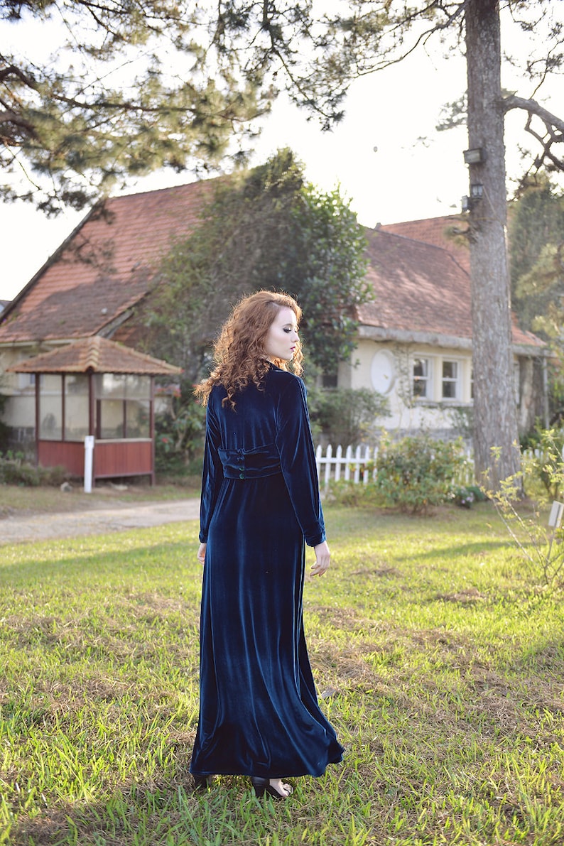 May include: A long, dark teal velvet dress with long sleeves and a cinched waist. The dress is worn outdoors in a grassy area with a house in the background. The dress has a high collar and a flowing skirt.