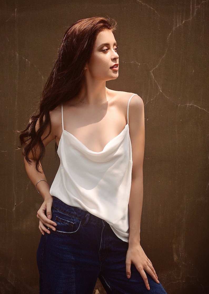 May include: A woman wearing a white cowl neck tank top and blue jeans. She is standing in front of a brown wall.