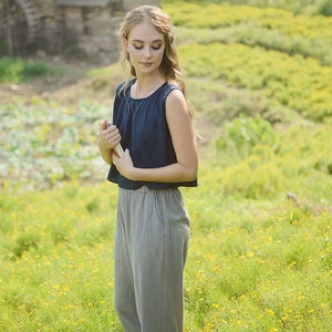 May include: A woman in a blue sleeveless top and gray pants stands in a field of yellow wildflowers. She is holding a book in her hands and looking down at it.