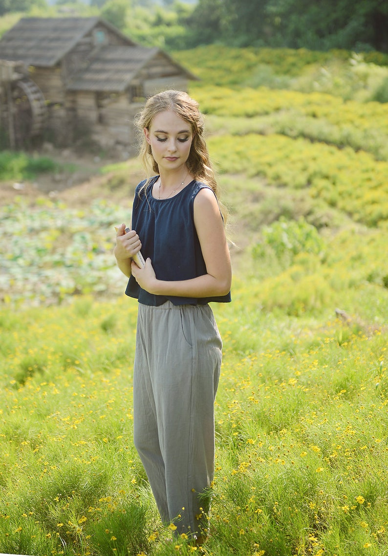 May include: A woman in a blue sleeveless top and gray pants stands in a field of yellow wildflowers. She is holding a book in her hands and looking down at it.