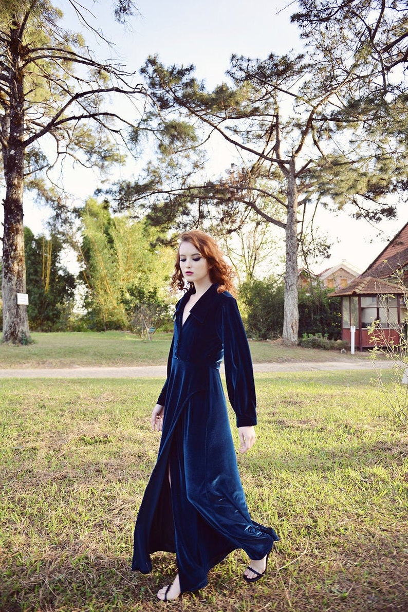 May include: A long, dark teal velvet dress with long sleeves and a high slit. The dress is buttoned at the waist and falls to the ankles. The person is wearing black sandals and walking on grass.