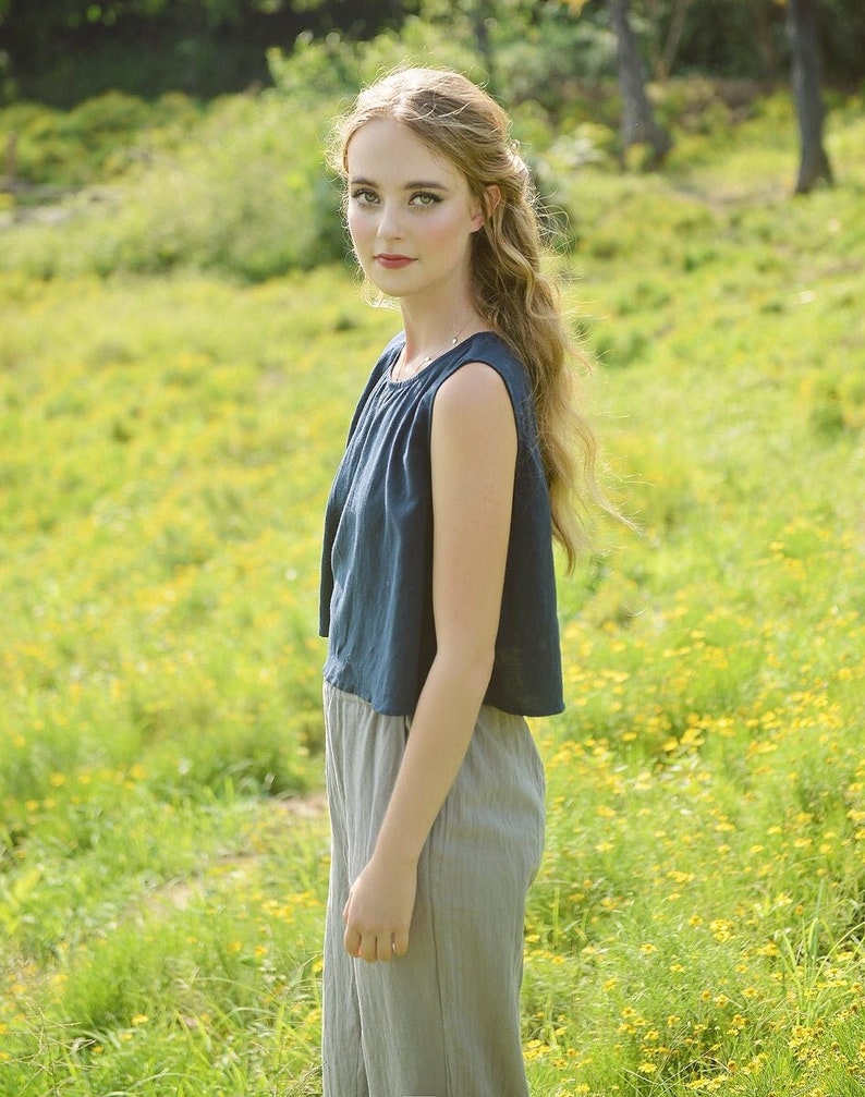 May include: A woman with long blonde hair wearing a dark blue sleeveless top and light gray pants stands in a field of yellow wildflowers.