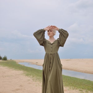 May include: A woman wearing a long, olive green, button-down dress with puffed sleeves stands on a sandy beach with a small stream of water in the background.