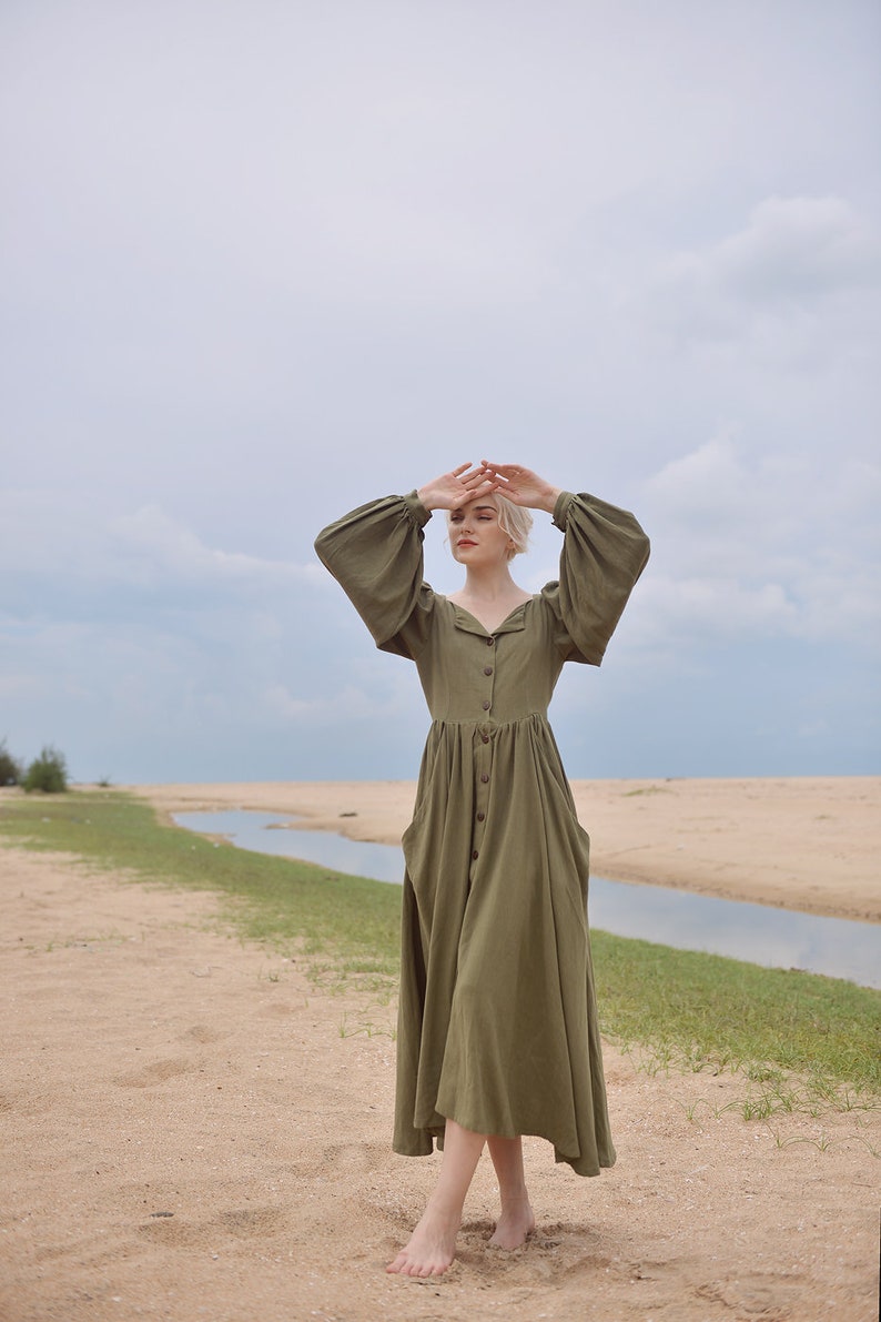 May include: A woman wearing a long, olive green, button-down dress with puffed sleeves stands on a sandy beach with a small stream of water in the background.