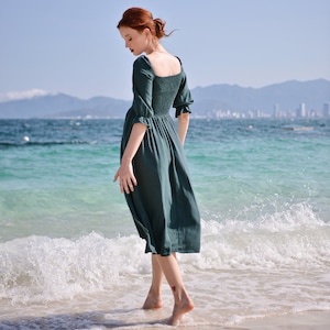 May include: A woman in a teal green dress walks on a sandy beach with turquoise water and white waves.