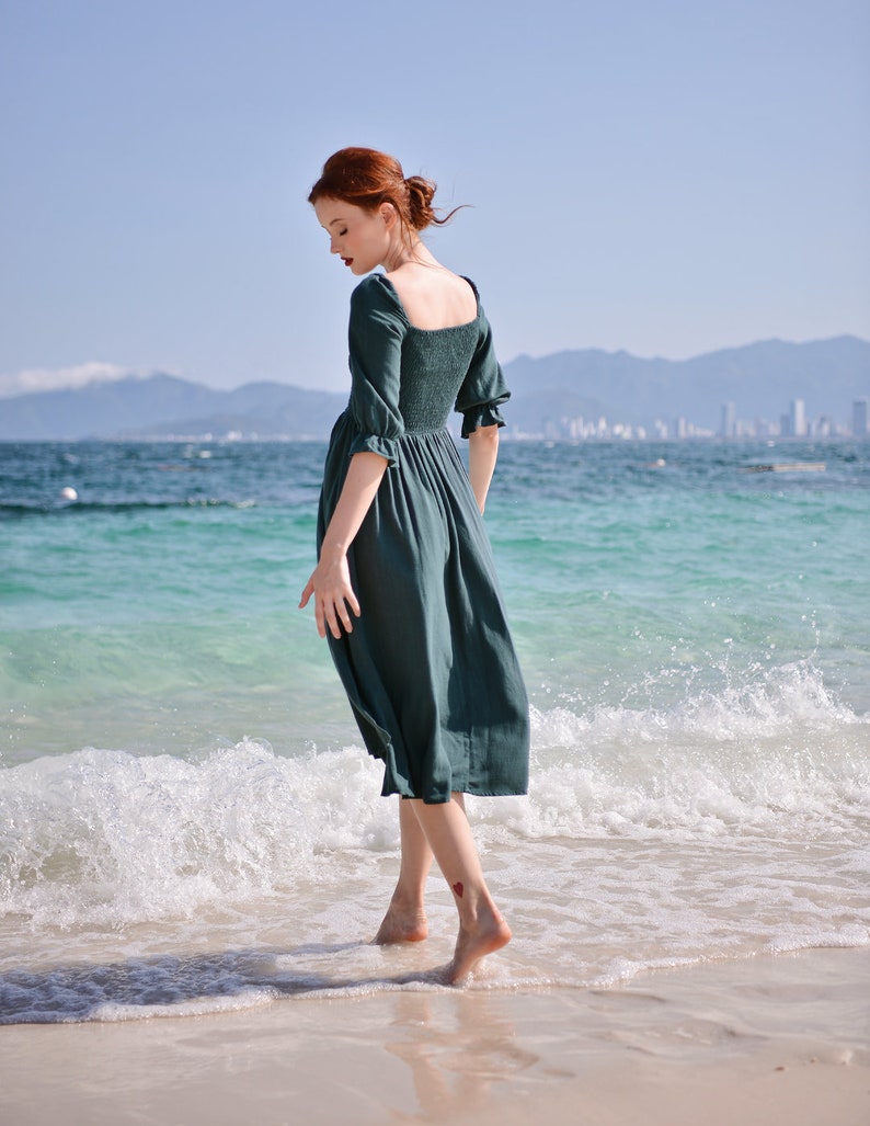 May include: A woman in a teal green dress walks on a sandy beach with turquoise water and white waves.