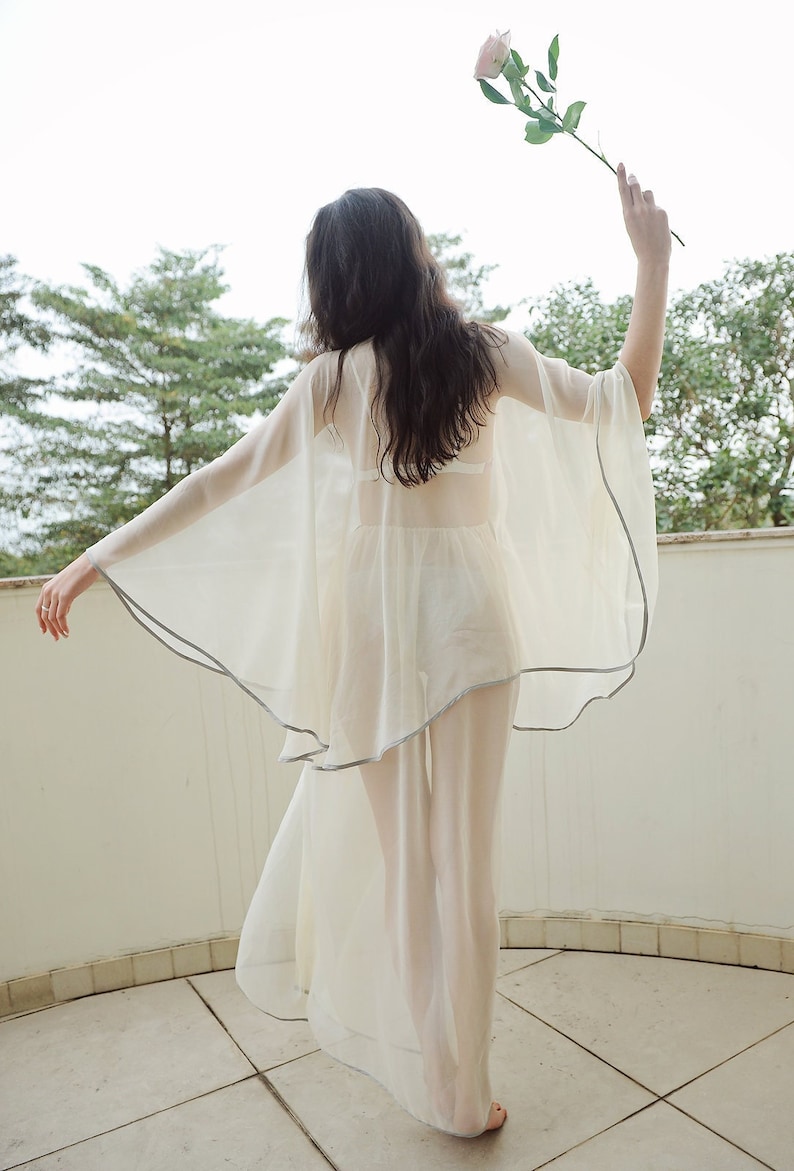 May include: A woman wearing a sheer white gown with a delicate gray trim. The gown is flowing and billowing in the breeze. She is holding a single pink rose in her hand.