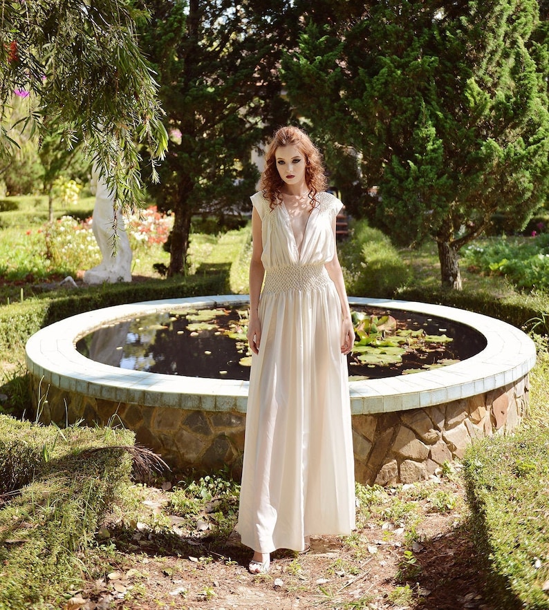 May include: A cream-colored, floor-length dress with a deep V-neck and a fitted waist. The dress features a flowing skirt and cap sleeves. The woman is standing in a garden with a pond in the background.