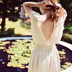 May include: A cream-colored dress with a low back and a fitted waist. The dress has short sleeves and a flowing skirt. The person is standing near a pond with lily pads. The person is holding their hair up.