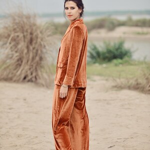 May include: A woman wearing a rust-colored velvet pantsuit. The pantsuit has a blazer with a single button closure and wide-leg pants.