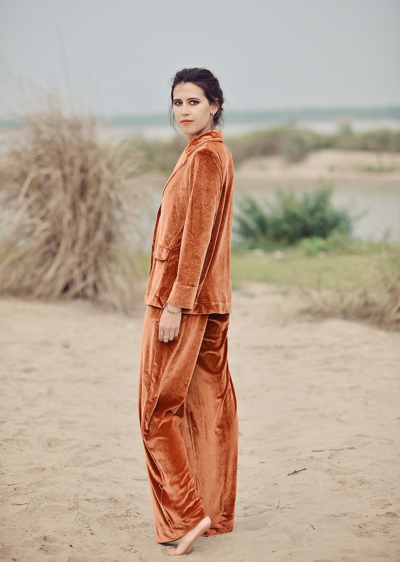 May include: A woman wearing a rust-colored velvet pantsuit. The pantsuit has a blazer with a single button closure and wide-leg pants.
