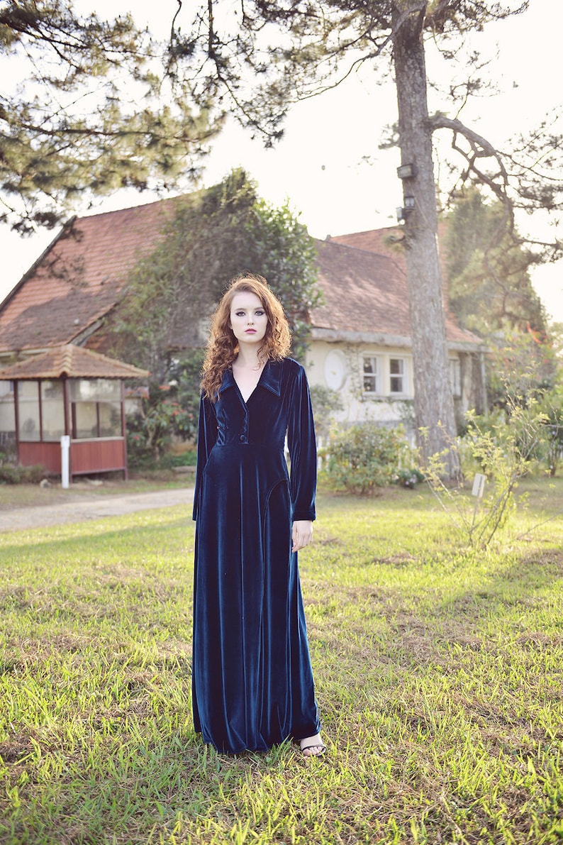 May include: A long, dark teal velvet dress with long sleeves and a collared neckline. The dress is worn outdoors in a grassy area, with a house and trees in the background. The dress has a button closure and a flowing skirt.