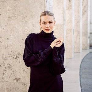 May include: A woman wearing a black turtleneck dress with long sleeves and a ribbed texture. She is standing in front of a white wall with columns.