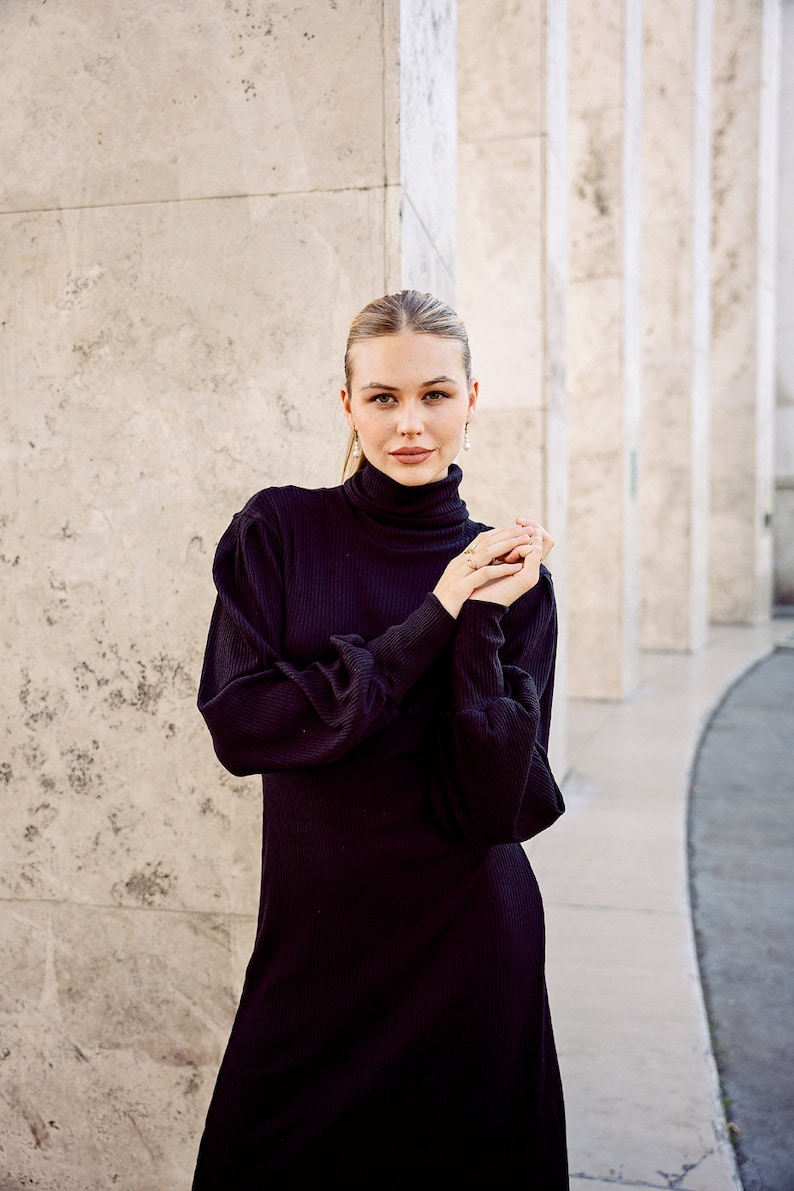 May include: A woman wearing a black turtleneck dress with long sleeves and a ribbed texture. She is standing in front of a white wall with columns.