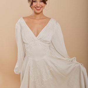 Op de afbeelding: Een witte, lange, V-hals jurk met een zwierige rok en delicate kantdetails.