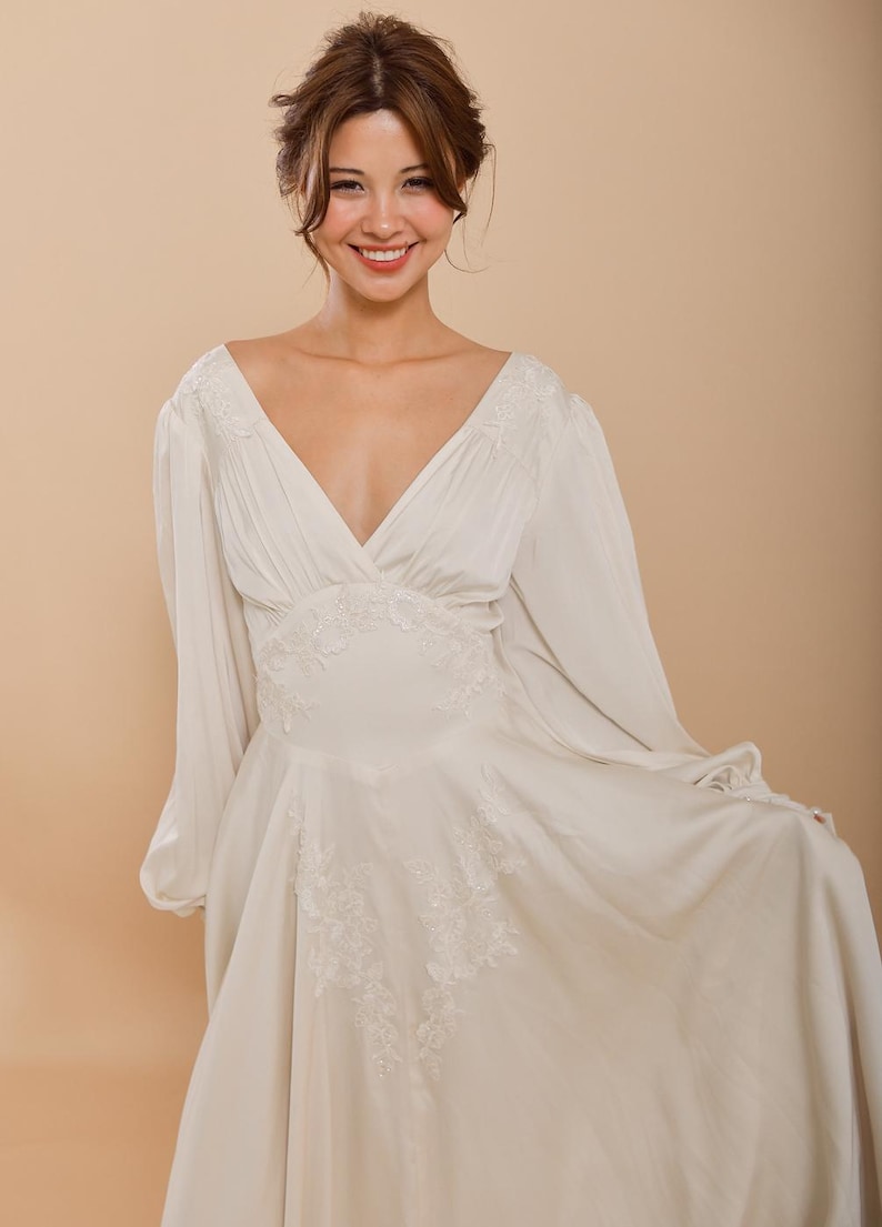 Op de afbeelding: Een witte, lange, V-hals jurk met een zwierige rok en delicate kantdetails.