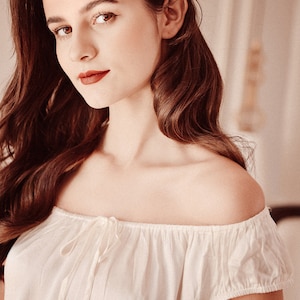 May include: A woman with long brown hair wearing a white off-the-shoulder top with a bow detail at the neckline.