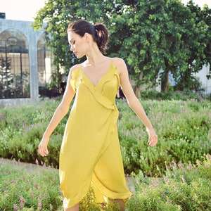 May include: A woman wearing a yellow wrap dress with spaghetti straps and a ruffled neckline. The dress is knee-length and made of a flowing fabric. The background features a garden with green plants and purple flowers.