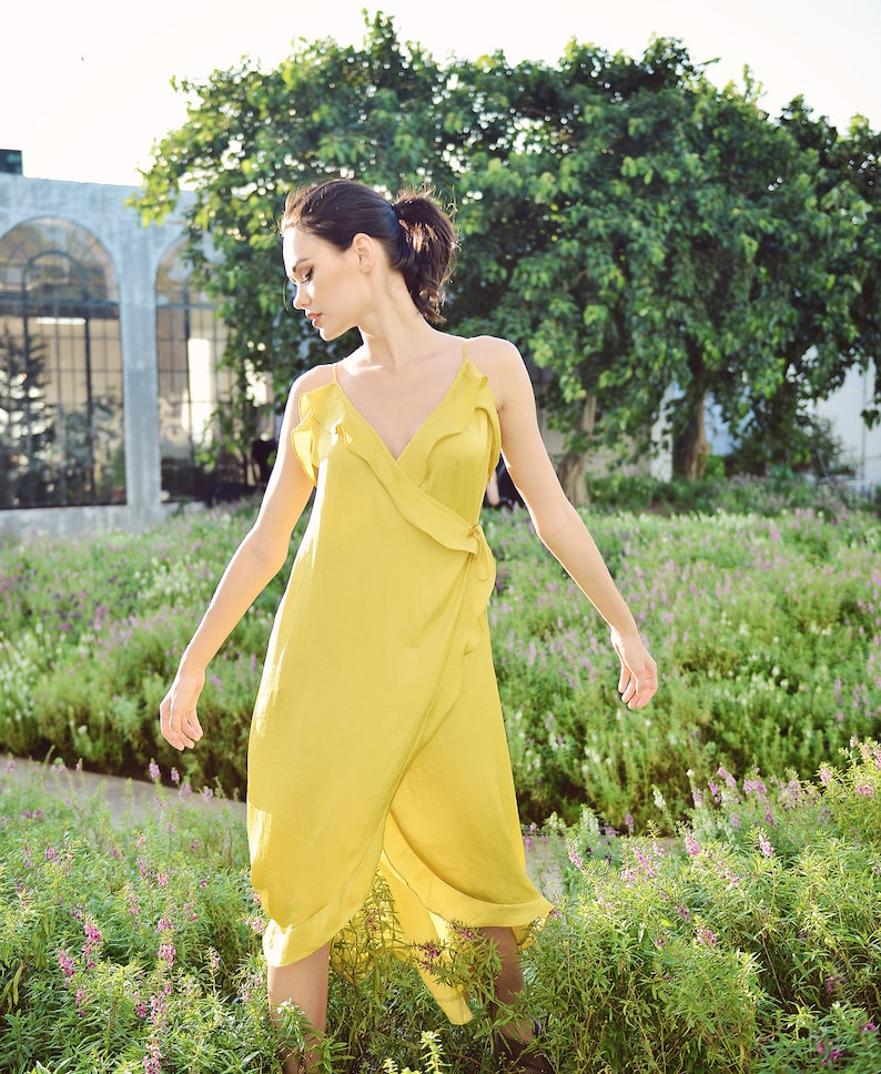 May include: A woman wearing a yellow wrap dress with spaghetti straps and a ruffled neckline. The dress is knee-length and made of a flowing fabric. The background features a garden with green plants and purple flowers.