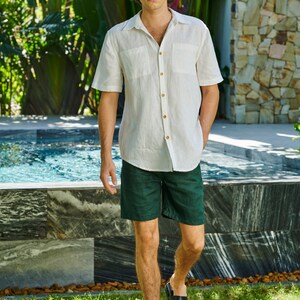 May include: A man wearing a white linen shirt with a button-down collar and a pocket on the left side. He is also wearing dark green linen shorts and black sandals. He is standing in front of a swimming pool.
