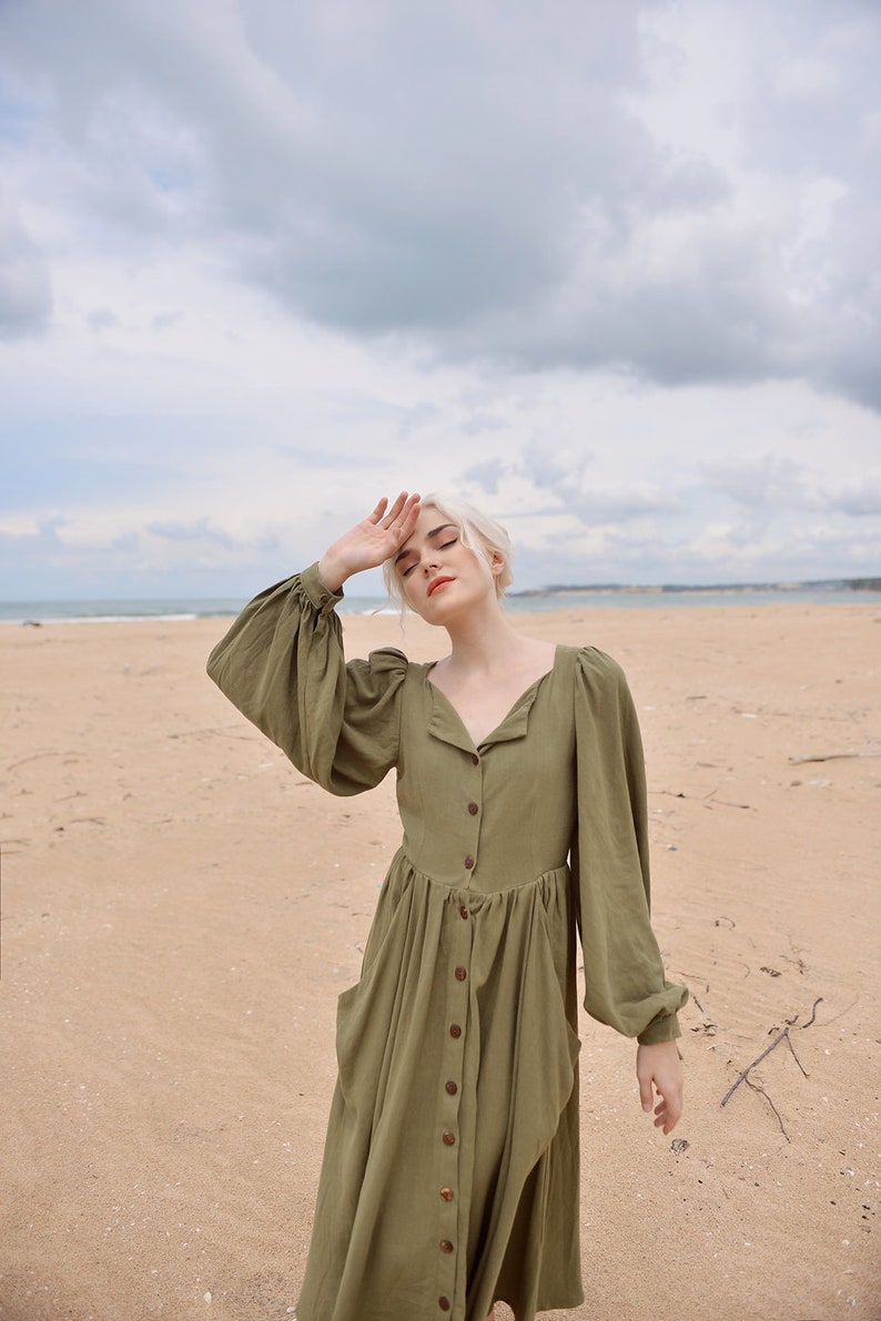 May include: A woman with blonde hair wearing a long, olive green, button-down dress with puffed sleeves. She is standing on a sandy beach with the ocean in the background.