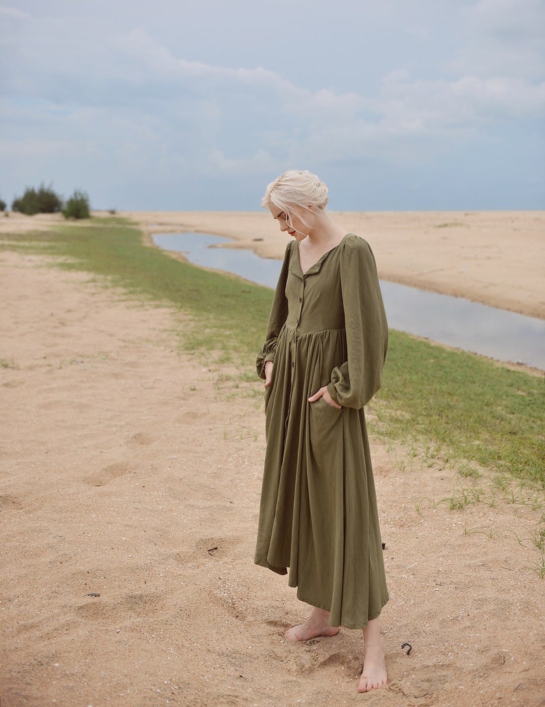 May include: A woman wearing a long, olive green, button-down dress with long sleeves and pockets. She is standing on a sandy beach with a small stream of water in the background.